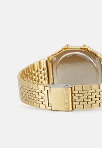 Montre-bracelet en métal doré avec un design de bracelet à maillons. Fermeture à boucle avec logo gravé. Fond de boîtier octagonal avec une surface lisse et brillante.
