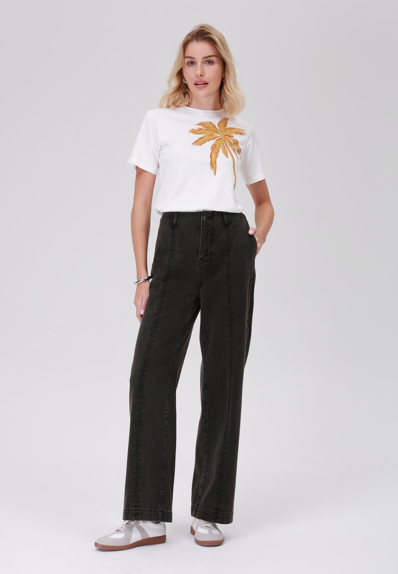 Witte t-shirt met een gouden palmbladprint, gecombineerd met hooggehaakte, rechte zwarte jeans. Model draagt sneakers in neutrale kleuren.