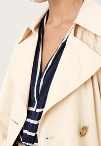 Gabardina beige con solapas grandes y detalle de botones dorados, sobrepuesta a una prenda azul marino con rayas blancas. Tejido liso y estructurado.