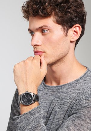 Junger Mann mit lockigem Haar, der einen grauen Pullover trägt, stützt das Kinn auf die Faust und zeigt eine schwarze und silberne Metallarmbanduhr.