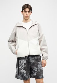 Νέος άνδρας που φοράει ανοιχτό γκρι και λευκό γουίντμπρέικερ με κουκούλα και σορτς με αφηρημένο μοτίβο σε μαύρο και γκρι, στέκεται μπροστά σε ένα απλό φόντο.