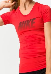 Punainen Nike-urheilupaita, jossa on lyhyet hihat, monipuolinen muotoilu ja iso musta logo edessä. Sileä kangas ja V-kaula-aukko.