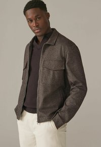 Ruda herringbone tekstūros zip-up striukė su dviem priekinėmis kišenėmis, apsivilkta virš tamsaus megztinio. Lygus paviršius, laisvas siluetas ir minimalus dizainas.