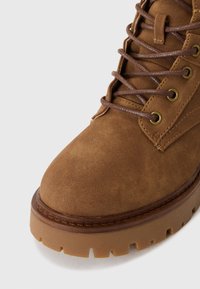 Bottines en suede marron avec un bout arrondi, œillets en métal, lacets marron et une semelle en caoutchouc texturée avec un motif à crampons.