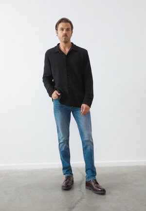 Camisa de botones negra con un patrón texturizado, combinada con jeans de color azul claro y zapatos marrones de cordones, de pie sobre un suelo de concreto.