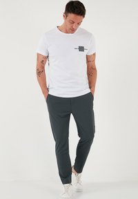 Witte korte mouwen T-shirt met een zwart grafisch zakontwerp, gecombineerd met donkergrijze slim-fit broek en witte sneakers, in staande pose.