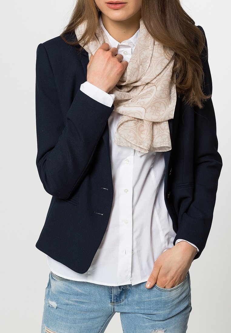 Femme portant un blazer bleu marine, une chemise blanche boutonnée, une écharpe beige à motifs et un jean bleu clair usé, une main dans la poche et l'autre tenant l’écharpe.