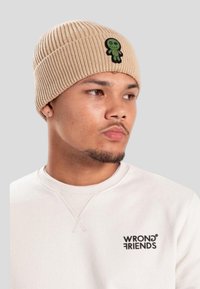 Młody mężczyzna w beżowej dzianinowej czapce z zielonym naszywką w kształcie lalki oraz białej bluzie z logo "WRONG FRIENDS", patrzący w prawo na szarym tle.