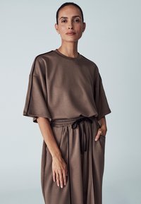 Brown, oversized top with short sleeves, made from smooth fabric. Drawstring waistband on matching relaxed-fit pants, featuring side pockets.
