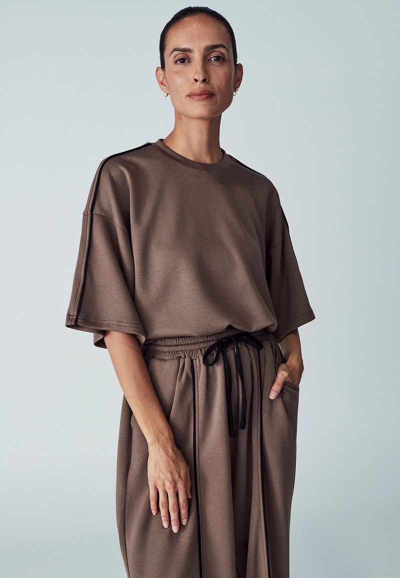 Brown, oversized top with short sleeves, made from smooth fabric. Drawstring waistband on matching relaxed-fit pants, featuring side pockets.