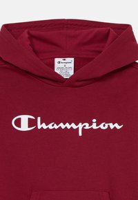 Maroon huppari, joka on valmistettu puuvillasekoitteesta, ja jossa on etupuolella kengurutasku sekä valkoinen "Champion"-logo. Hupun sisävuori on samaa maroonia.
