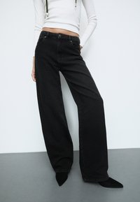 Donna che indossa jeans a vita alta neri a gamba larga con scintillii sottili, una maglietta bianca a maniche lunghe e corta, e scarpe nere con punta affilata su uno sfondo semplice.