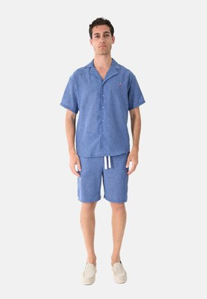 Giovane uomo in piedi che indossa una camicia azzurra a maniche corte con bottoni e pantaloncini coordinati con lacci bianchi e scarpe beige slip-on.