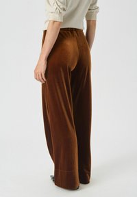 Pantaloni largi din catifea maro, cu talie înaltă și o strălucire subtilă, având o textură netedă și o croială lejeră.