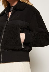 Czarna kurtka bomberka z teksturą fleecową i gładkimi panelami materiałowymi. Posiada kieszenie zapinane na zamek oraz ściągacze na mankietach. Dyskretna marka na klatce piersiowej.