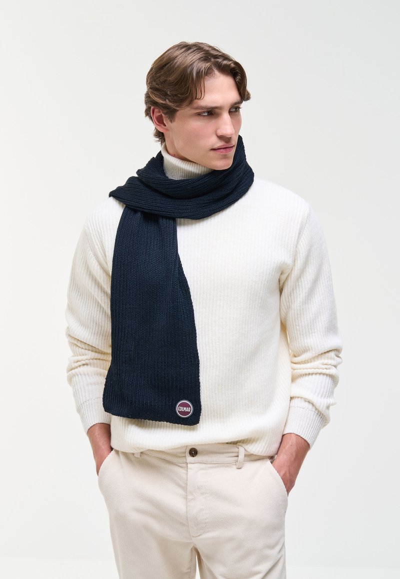 Jeune homme portant un pull côtelé blanc et un pantalon beige avec une écharpe tricotée bleu foncé enroulée autour du cou.