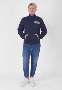 Giacca zip-up navy con colletto alto e tasca sul petto grigia. Indossata con jeans azzurri chiari e scarpe da ginnastica bianche. Tessuto liscio, design aderente.