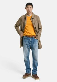 Beiger Trenchcoat, leuchtend orangener Pullover, hellblaue Jeans und braune Schuhe. Bemerkenswerte Details: strukturierter Gürtel, gestapelter Look, lässiger Stil.