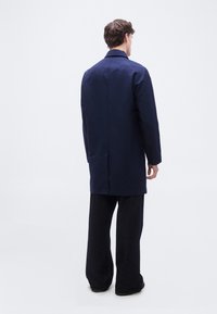 Άνδρας στέκεται γυρισμένος με την πλάτη, φορώντας ένα μπλε παλτό μέχρι το γόνατο και φαρδιά μαύρη παντελόνα, πάνω σε λευκό φόντο.