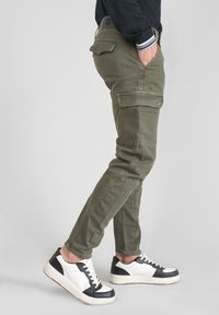 Osoba w oliwkowych spodniach cargo i czarno-białych trampkach, stojąca z jedną ręką w kieszeni na neutralnym tle.