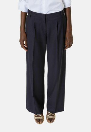 Pantalones anchos color azul marino oscuro combinados con camisa blanca y sandalias doradas de tiras, en una persona de pie frente a un fondo liso.