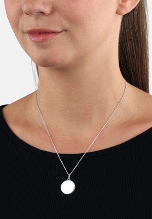 Collana con ciondolo circolare in argento con una finitura liscia, sospesa su una sottile catena, indossata sopra un top nero a coste.