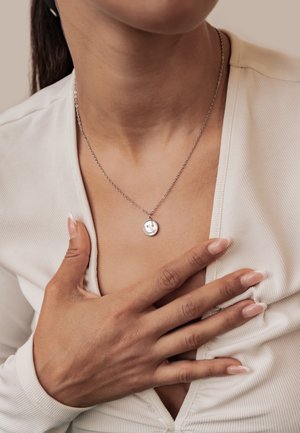 Woman with manicured nails wearing a silver chain necklace with a round pendant over a white ribbed top.
