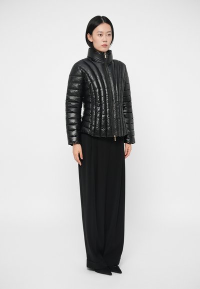 Doudoune noire avec un col haut, présentant un design matelassé et une texture brillante. Fermeture à glissière à l'avant. Modèle portant un pantalon noir à jambes larges.