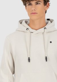 Lichtbeige hoodie met een trekkoordcapuchon, zwarte hardwareaccenten en een voorkantzak. Zachte textuur met een subtiele glans en minimale ontwerpdetaillering.