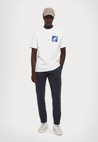 Vit kortärmad t-shirt med blå grafisk design, i kombination med mörkgrå slim-fit byxor och vita sneakers. Inkluderar en beige keps.