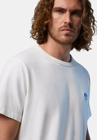 T-shirt bianco in cotone con scollo rotondo e maniche corte. Presenta un piccolo logo circolare blu sul petto sinistro. Texture liscia.