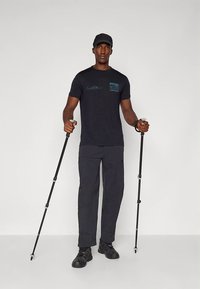 Svart T-skjorte med grafikk på brystet, kombinert med mørkegrå cargo-bukser og solide svarte sko, holder to vandrestaver.