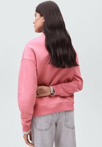 Žena s dlouhými tmavými vlasy v růžové mikině a světlých šedých džínech, stojící s jednou rukou za zády před jednobarevným pozadím.