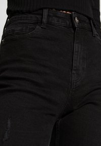 Jeans de mezclilla negra con un diseño ajustado, bolsillos visibles, cierre de botón y detalles desgastados en la parte inferior de la pierna.