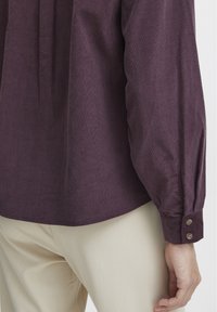 Burgundy long-sleeved shirt with a textured finish, featuring a buttoned cuff and pleats along the back. Paired with light beige pants.