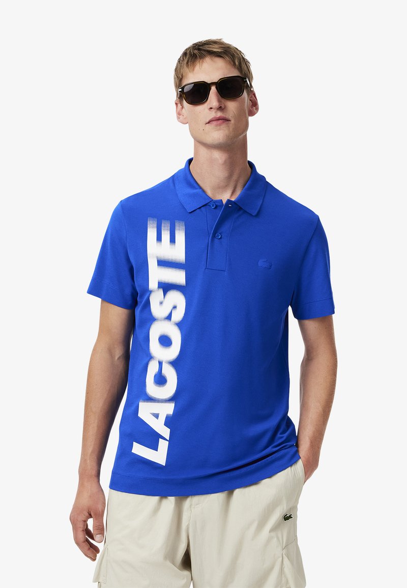 Sininen puuvillapaita, jossa on lyhyet hihat. Vasemmalla puolella on suuri valkoinen "LACOSTE"-logo ja rinnassa pieni logo.