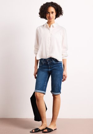 Kobieta nosząca podwinięte dżinsowe szorty, białą koszulę na guziki, czarne sandały, trzymająca czarną torbę na jednolitym tle.