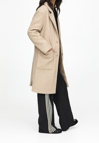 Abrigo de lana beige con collar de pico, bolsillos laterales, cierre de un solo botón y una silueta estructurada, combinado con pantalones negros con rayas blancas.