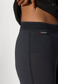 Melnas legingas no Polartec auduma, ar teksturētu jostu un smalku svītrojumu sānos. Tuvplāns, kas izceļ materiālu.
