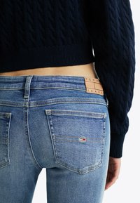 Jeans de denim azul con un ligero desvanecimiento, que presentan una cinturilla texturizada y una etiqueta de cuero marrón con la inscripción "TOMMY JEANS" y un pequeño logotipo en la parte posterior.