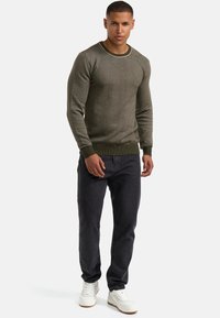 Pullover tricotat de culoare verde măslin, cu guler rotund și manșete ribbenate, asortat cu blugi gri închis și adidași albi.