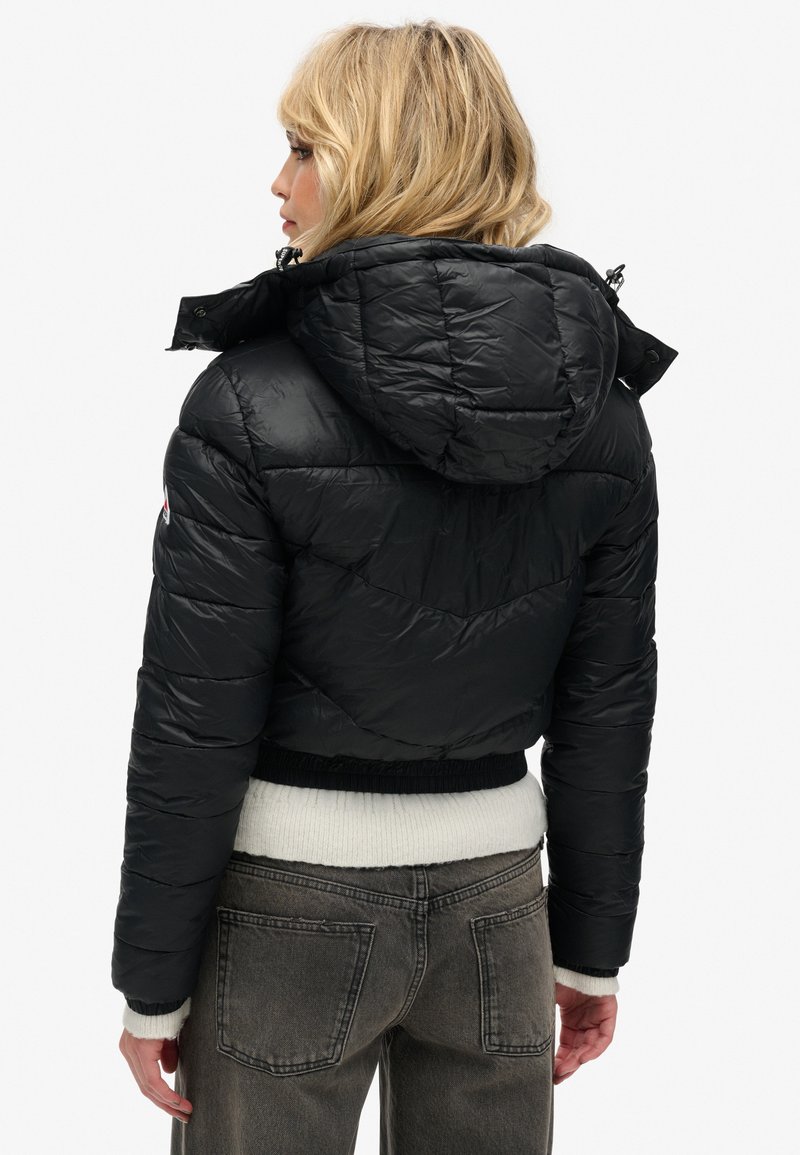 Giubbotto piumino nero con cappuccio, caratterizzato da texture trapuntata, polsini elasticizzati e design corto. Visualizzato dal retro, mostrando i dettagli del materiale.