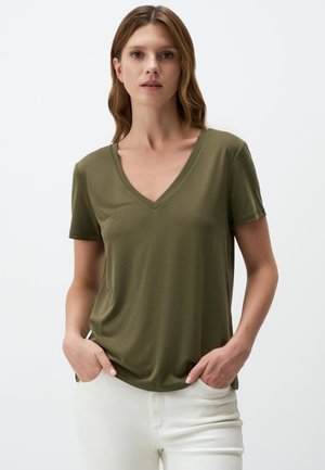 T-shirt vert olive à manches courtes et col en V, fabriqué en tissu doux, avec une coupe décontractée et de légentes fentes sur les côtés. Porté avec un jean blanc.