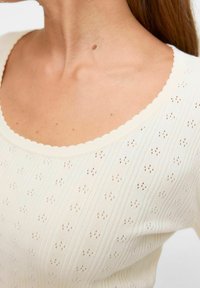 Lichtcrème, gebreide top met korte mouwen en een scalloped halslijn, voorzien van een textuurpatroon van kleine gaten en verticale ribbels.