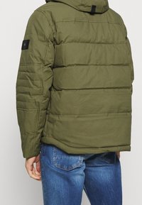 Uomo che indossa una giacca trapuntata verde oliva e jeans blu, visto da dietro con il braccio sinistro rilassato al suo fianco.