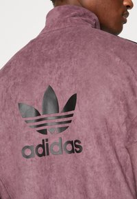 Lila mockaskinnsjacka med hög krage, med en svart adidas-logotyp och design med tre ränder på baksidan. Slät yta.