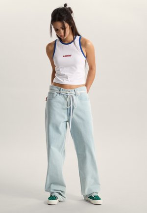 White sleeveless tank top with blue accents, paired with light blue oversized jeans and green checkered slip-on shoes.