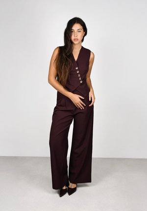 Mujer con cabello largo y oscuro que lleva un chaleco sin mangas de color púrpura oscuro con botones dorados y pantalones de pierna ancha a juego, posando frente a un fondo simple.