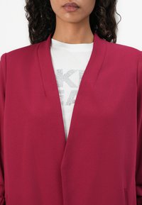 Blazer fuchsia avec un col châle et une ouverture devant. Porté par-dessus une chemise blanche avec des lettres argentées. Texture lisse, coupe ajustée.