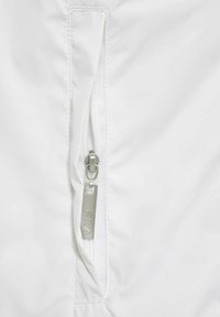 Valkoinen takki, jossa on vetoketjullinen tasku hopeisella vetoketjun vetimellä, teksturoitua kangasta ja hienovaraisia ompelupistoja.